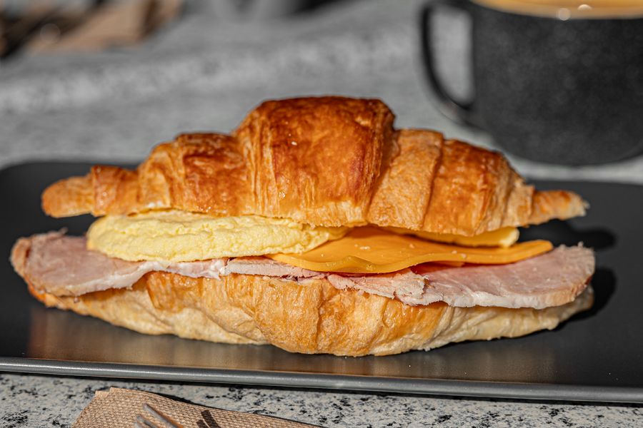 A breakfast croissant with ham, egg and cheese, with a cup of coffee behind it